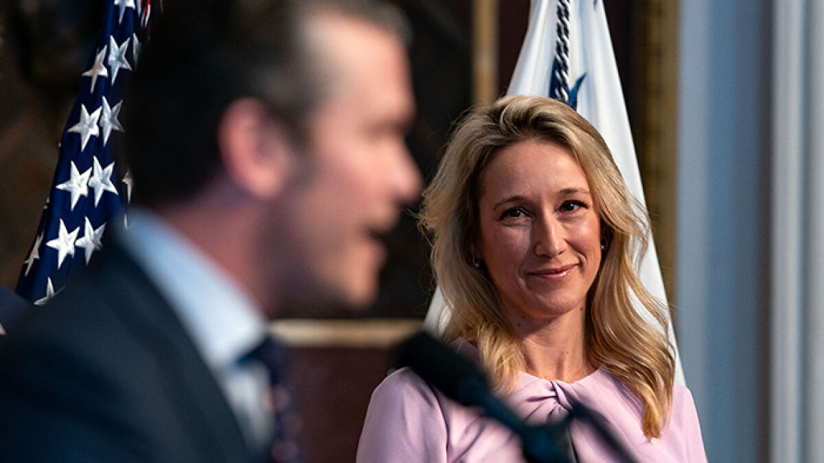 Jennifer Rauchet smiling during a Pentagon event with Pete Hegseth speaking in the foreground, American flags behind them.