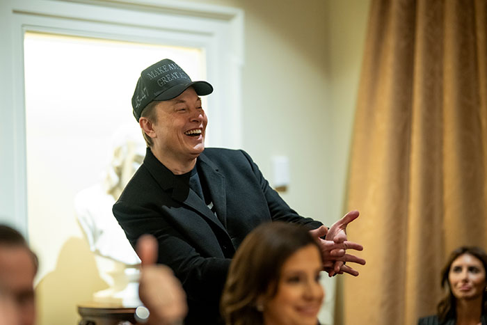 Man in a black jacket smiling, wearing a cap in a room, related to Tesla’s earnings and DOGE discussion.