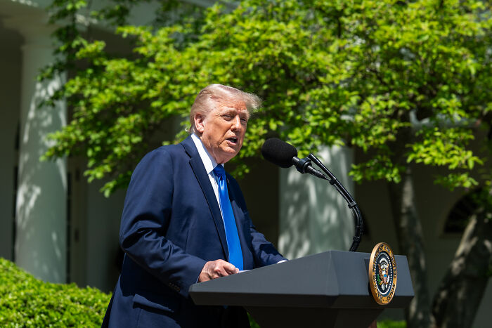Man speaking at a podium with presidential seal, outdoors with green trees, related to Mike Waltz ousted from post news.