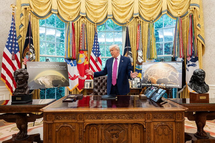Donald Trump discusses bold defense deal with Canada in the Oval Office, standing behind Resolute desk with flags and displays.