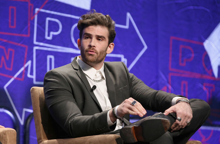 Popular left-wing Twitch streamer Hasan Piker speaking at an event, wearing a suit with a white shirt and chains.