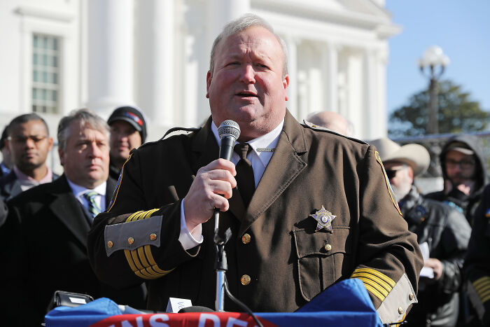 Sheriff in uniform speaking at outdoor event, representing law enforcement involved in badge bribery controversy.