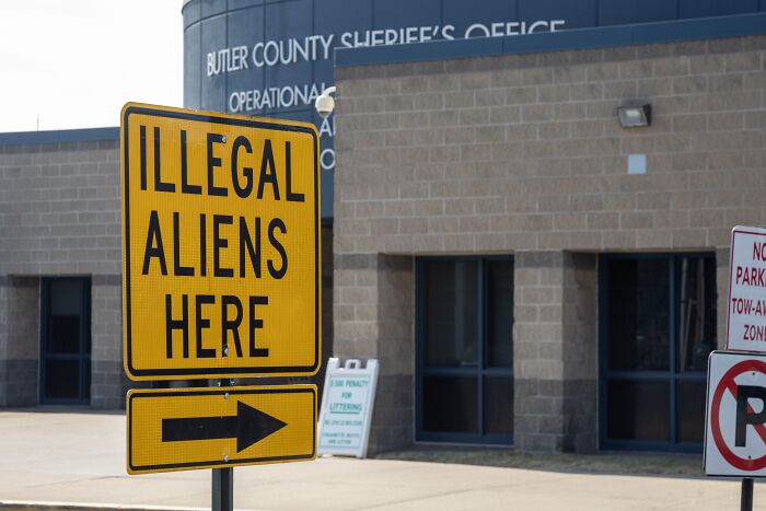 Yellow sign reading illegal aliens here with an arrow outside Butler County Sheriff’s Office referencing Asian migrants deported to African country.