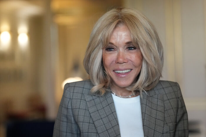 Blonde woman smiling in a checkered blazer indoors, related to bizarre scene involving Macron and his wife on plane argument.
