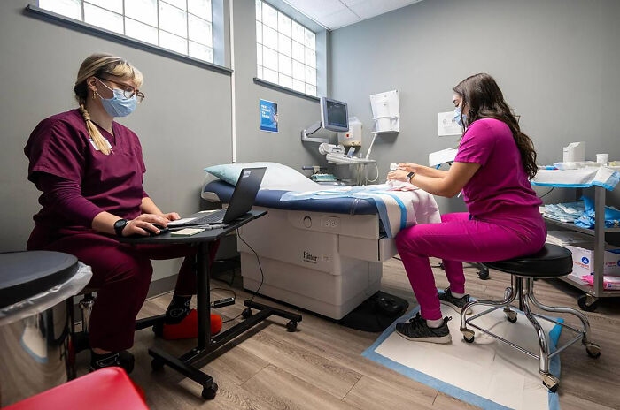 Two healthcare workers in a clinic setting, one using a laptop and the other preparing medical supplies, highlighting dystopian surveillance.