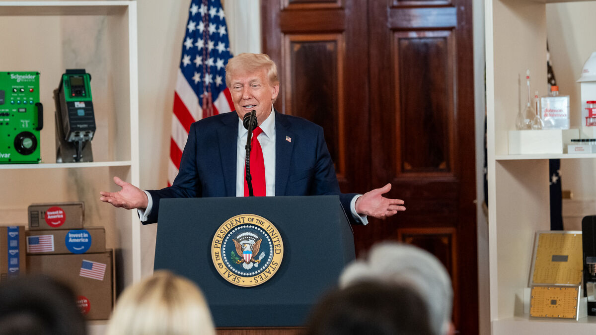 Donald Trump speaking at a podium with the presidential seal during a town hall event, audience visible in foreground.