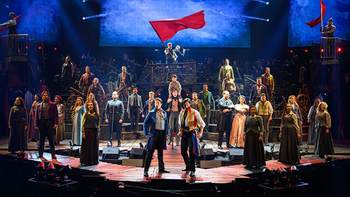 Les Misu00e9rables cast performing on stage with red flags and dramatic lighting during a live theater production.