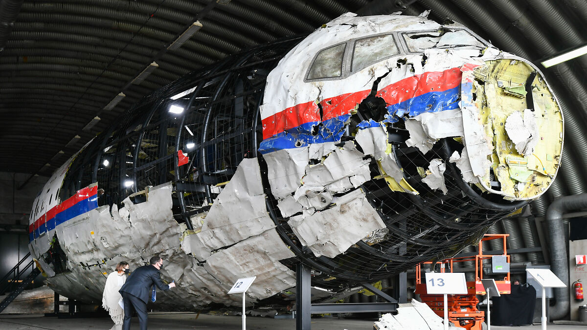 Wreckage of MH17 airplane on display in a hangar with people inspecting near aircraft parts and information boards.