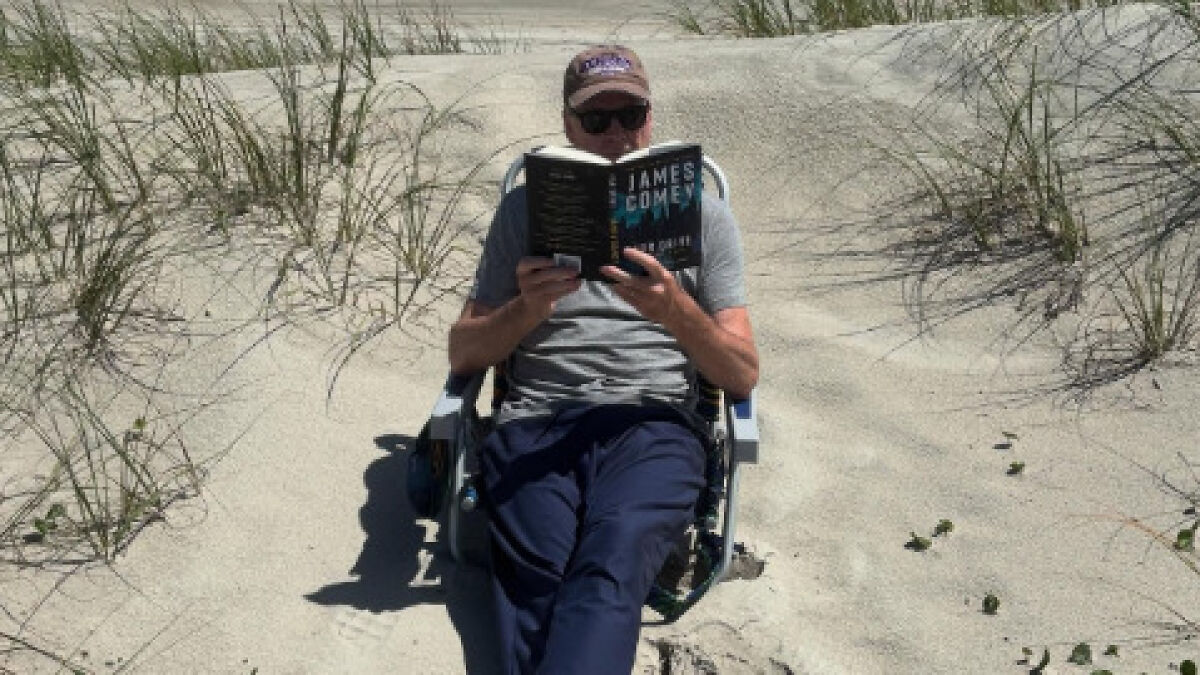 Man sitting on beach chair reading James Comey book in sandy area with sparse grass, casual clothing and cap on head.