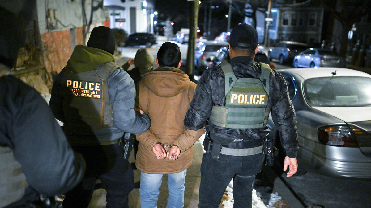 Two police officers escort a handcuffed migrant at night amid rising ICE detention center human rights concerns.