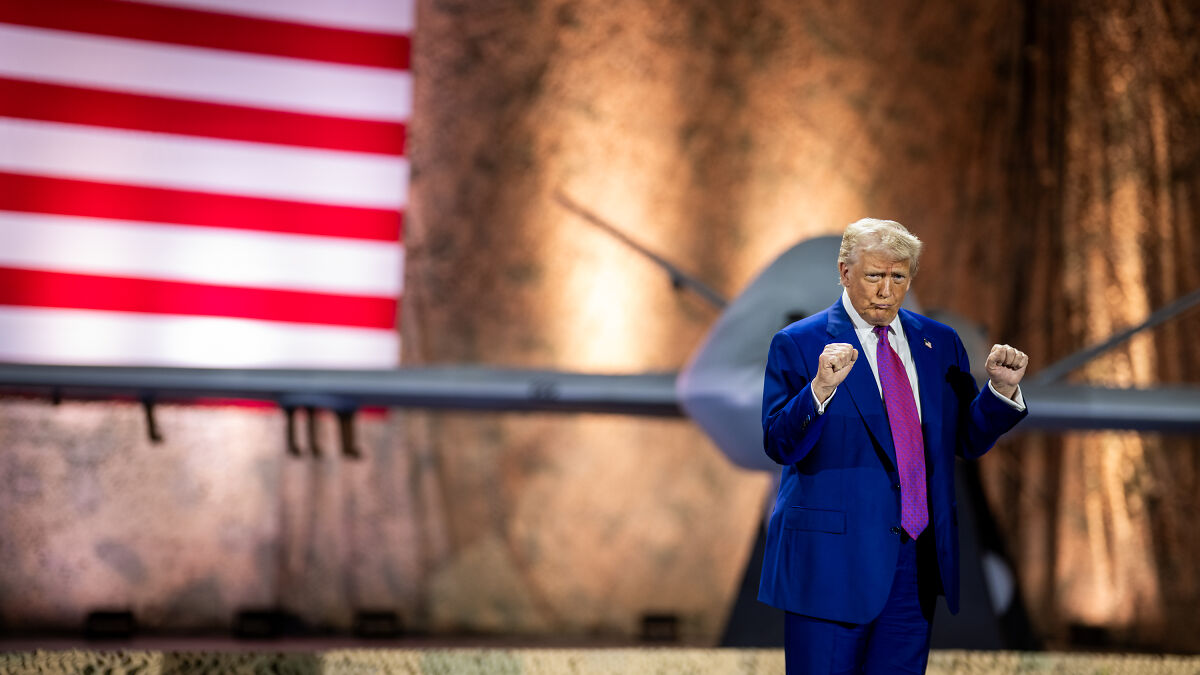 Donald Trump standing near a drone with an American flag in the background, raising his fists during a speech.