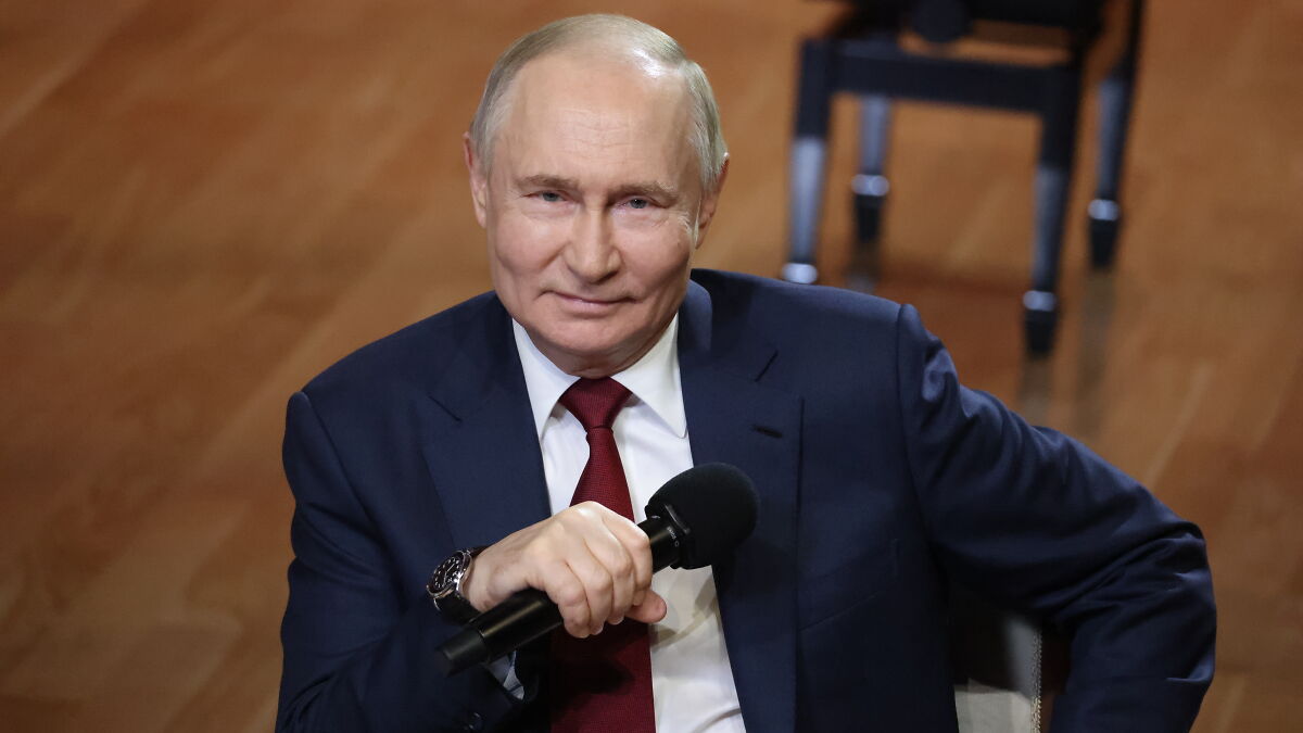 Vladimir Putin holding a microphone, wearing a dark suit and red tie, in a formal indoor setting.