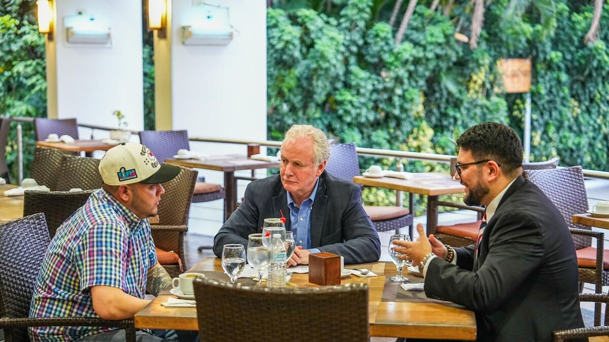 Three men in a serious discussion at a restaurant table about deportees and related political issues.