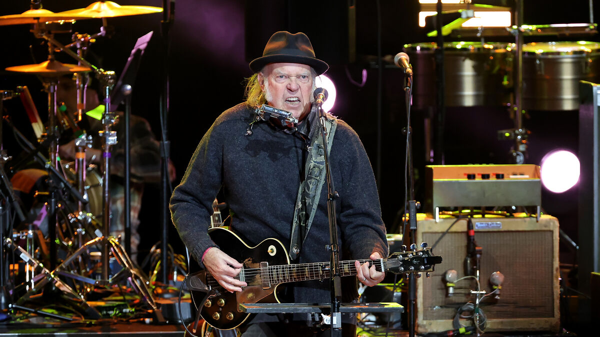 Neil Young performing live on stage with guitar and harmonica, highlighting musicians and political message.