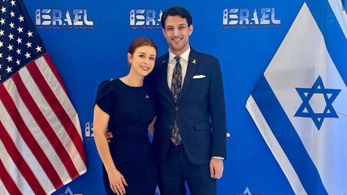 Israeli embassy couple posing together with US and Israeli flags at a formal event background.