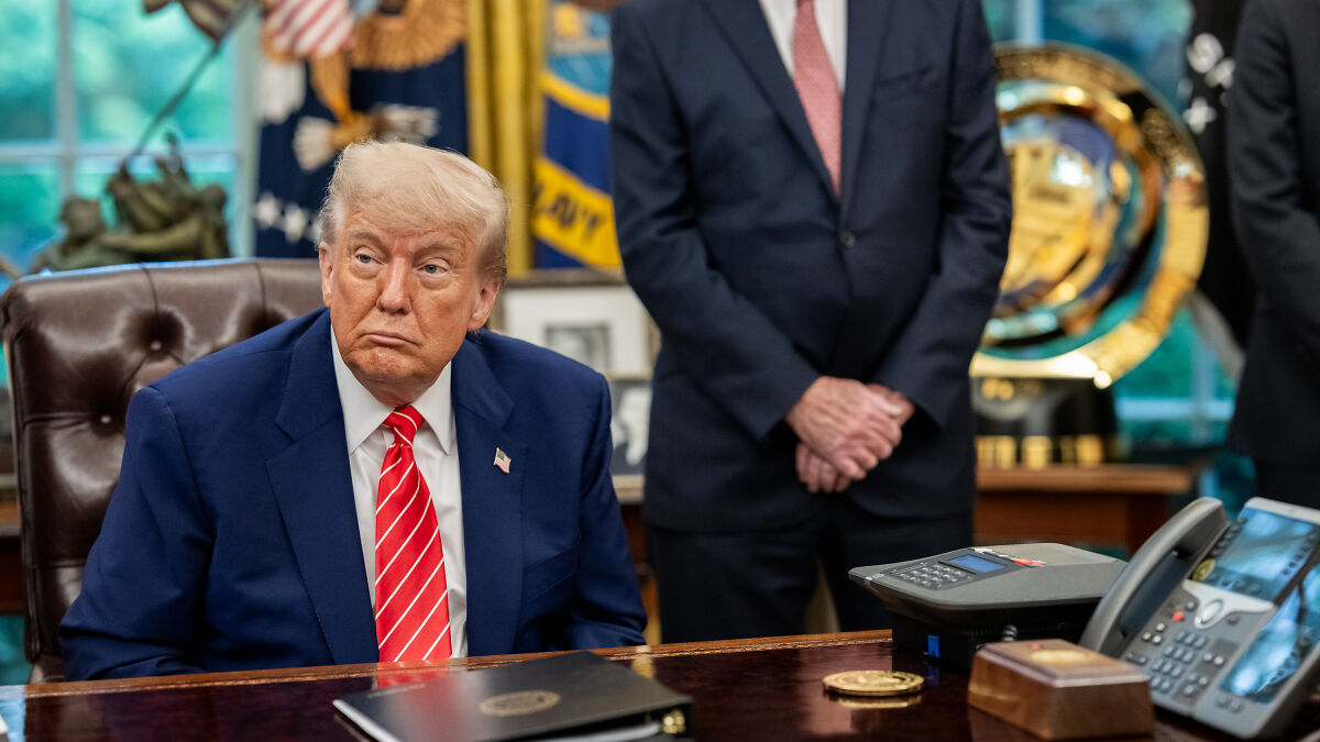 Donald Trump sitting at desk in Oval Office, wearing red striped tie, reacting to court ruling on tariffs.