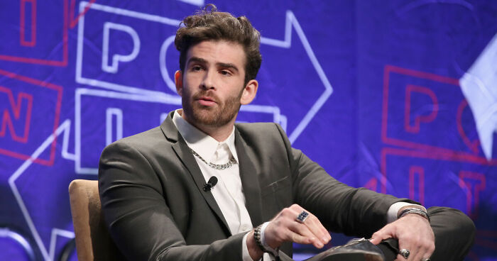 Hasan Piker in a suit speaking at an event, representing popular left-wing Twitch streamer detained at U.S. airport.