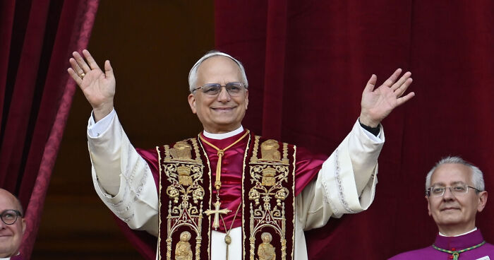 Pope Leo XIV wearing ornate religious robes and glasses, raising hands during a public event with clergy members nearby.
