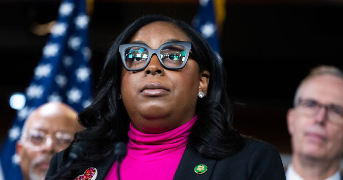 House Democrat speaking at a press event, with American flag in background, amid ICE facility visit and GOP arrest warnings.