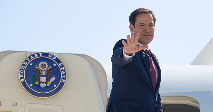 U.S. official waves near plane with Secretary of State seal amid student visa freeze and social media screening efforts.