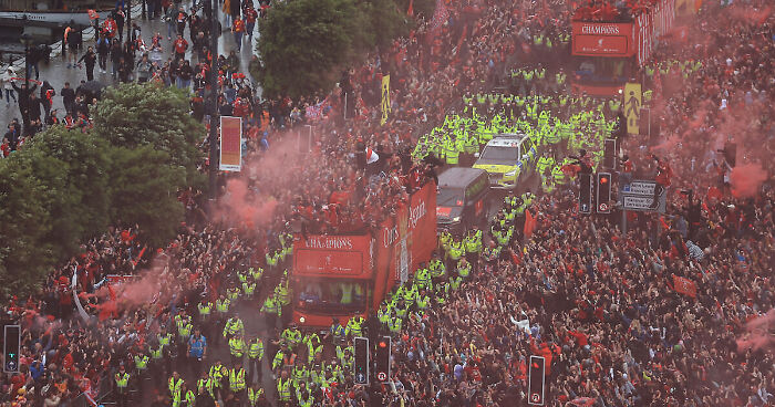 Crowds surrounding Liverpool FC parade buses with police presence amid fans vandalizing suspectu2019s car in chaotic scene.