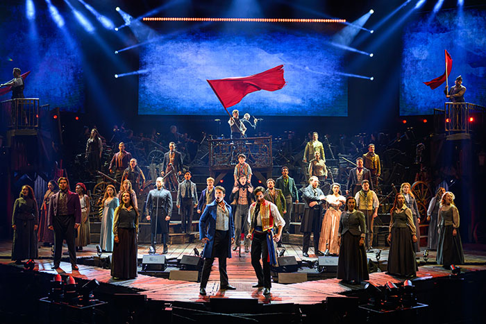 Les Misérables cast performing on stage holding red flags during a dramatic theater production.