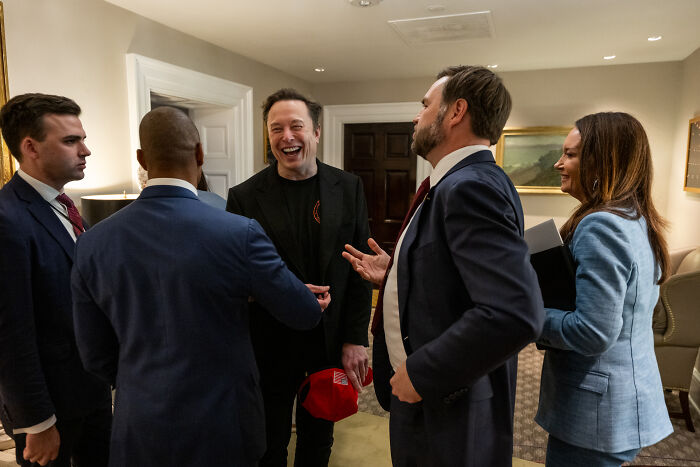 Group of professionals in a conversation indoors, with one man laughing, focusing on overweight and neurotic pal getting fat shot abroad.