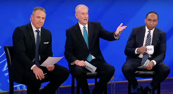 Three male news anchors in suits seated on stage during a town hall reacting to Trump’s first 100 days claims.