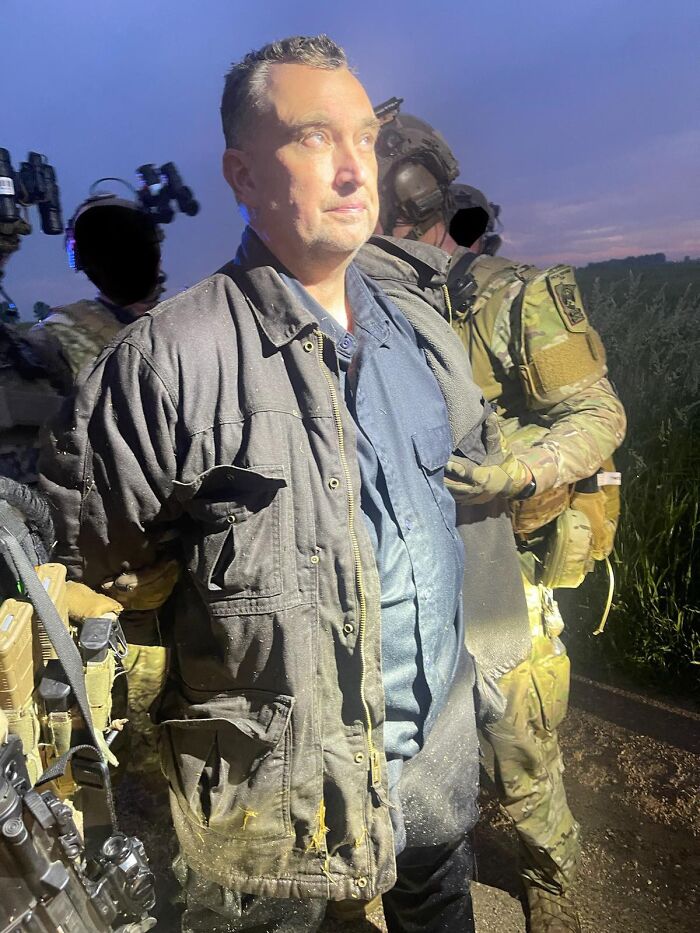 Alleged shooter in lawmakers attack wearing dark jacket, escorted by armed military personnel outdoors at dusk.