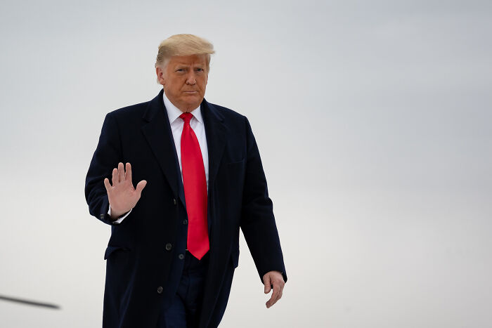 Donald Trump in dark coat and red tie, raising hand, symbolizing ICE ramping up immigration raids in blue cities directive.