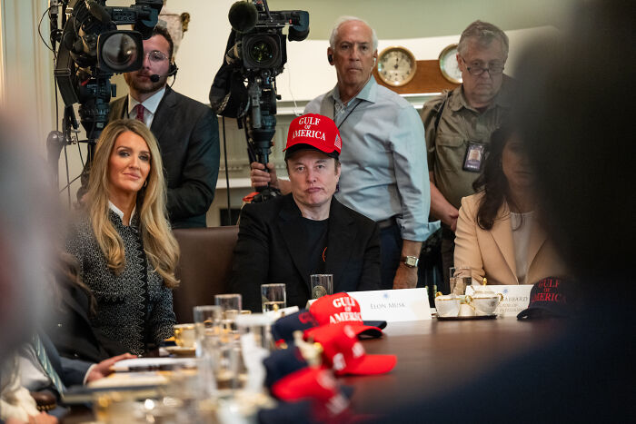 Elon Musk seated at a meeting table wearing a Gulf America hat, surrounded by attendees and media cameras.