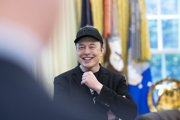 Elon Musk in a casual setting wearing a black cap and jacket, reacting during a discussion about foreign student ban.