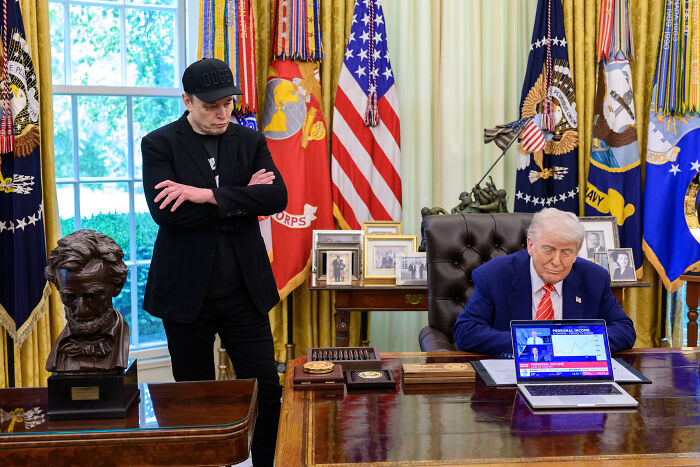 Elon Musk standing with arms crossed near Donald Trump seated at desk in the Oval Office with presidential flags.