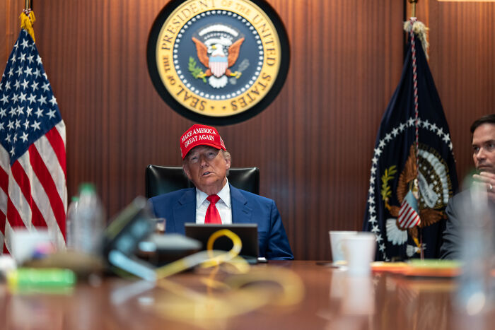 Donald Trump wearing a Make America Great Again hat, seated at a table with presidential flags and seal in background.