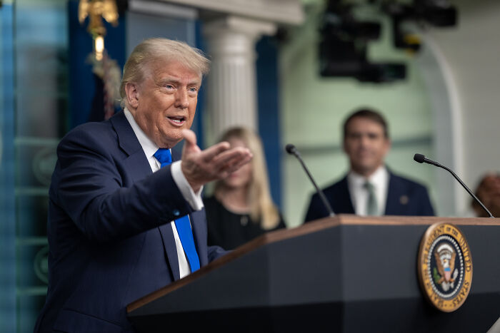Former President Trump speaking at a podium with presidential seal during a political event in the White House briefing room.