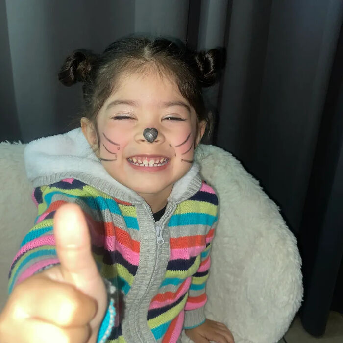 Smiling young girl with face paint and colorful sweater giving thumbs up, related to girl fatally shot by brother incident.