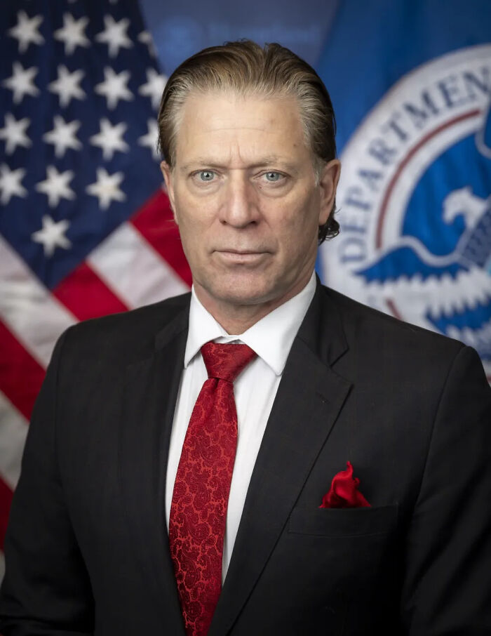 FEMA chief in a dark suit and red tie standing in front of US flag and Department of Homeland Security seal.