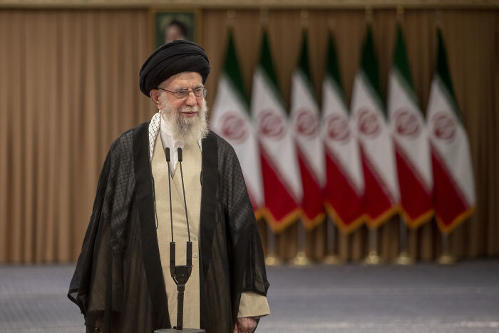 Iran’s Supreme Leader speaking at a podium with multiple Iranian flags in the background during a public statement.