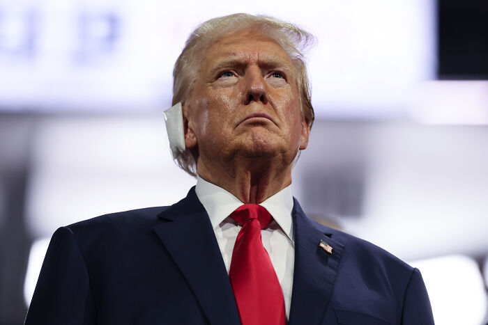 Donald Trump in a dark suit and red tie, looking serious during a public appearance related to assassination attempt footage.