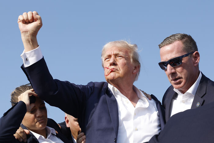 Former President Trump with blood on his face raising fist, escorted by security amid Trump assassination attempt footage.