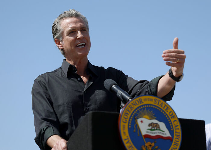 California Governor Gavin Newsom speaking at a podium outdoors with clear sky amid LA riots discussions.