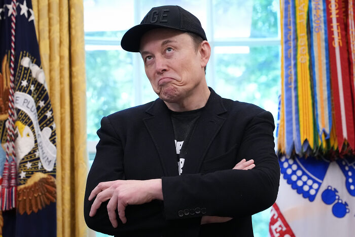 Elon Musk in a black cap and blazer with arms crossed, standing in an official room with flags and decorations.