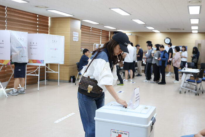 Woman casting vote in a busy polling station, highlighting Karoline Leavitt’s cringe binder shuffle over South Korea question.
