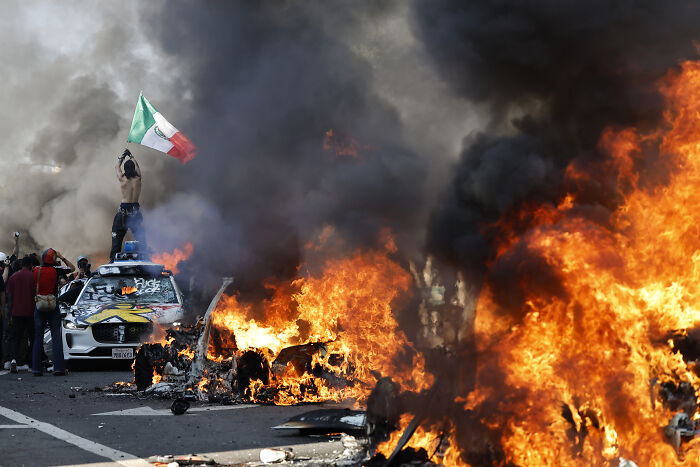 Violent ICE protestors amidst burning wreckage and thick smoke during chaotic street demonstrations in Florida.