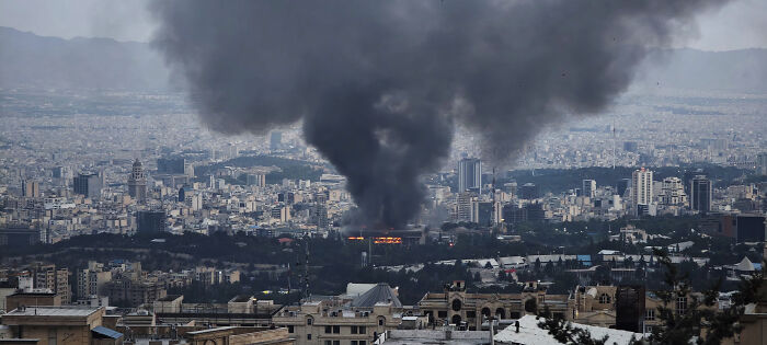 Thick black smoke rises over a city after an Israeli missile strike, with a female anchor running for life on Iran state TV.