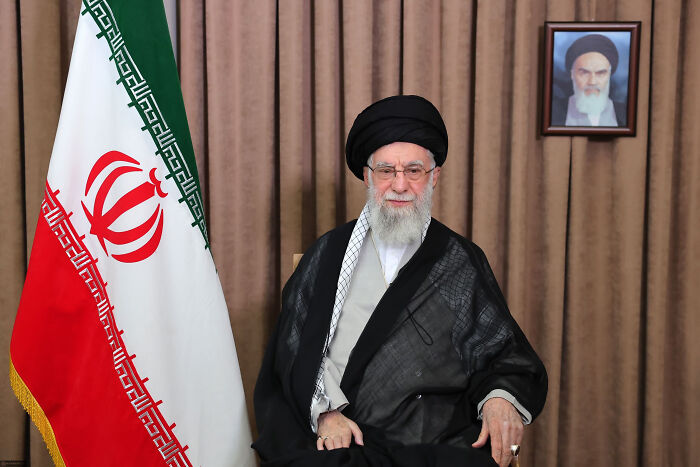 Iranian leader sitting beside Iran flag with a framed portrait in the background symbolizing Iran vows retaliation for nuclear strikes.