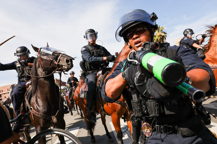 Police officers on horseback in Los Angeles, one aiming a launcher amid National Guard control during protests.