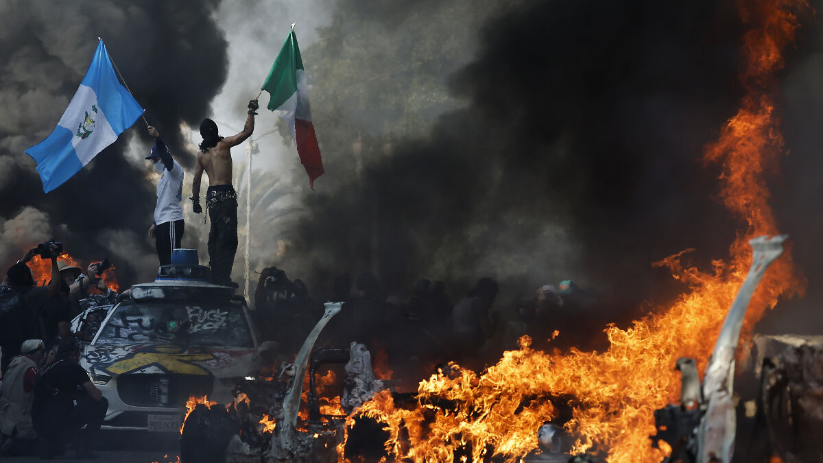 Protesters waving flags amid thick smoke and intense flames during riots in Los Angeles city streets.