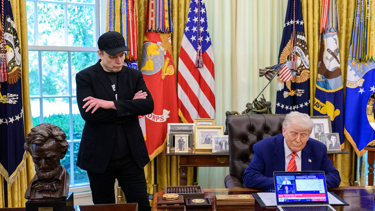Elon Musk standing with arms crossed near Donald Trump seated at desk with flags in background.