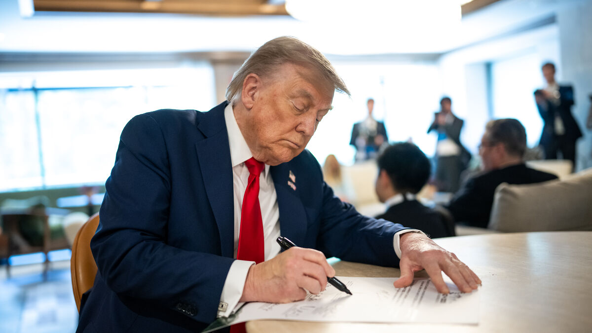 Donald Trump in a suit and red tie signing documents indoors amid blurred people, related to Iran attack plan approval.